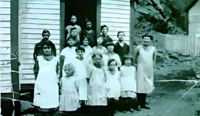 01 School kids c. 1929. Joe Lockyer is standing by the school on the left Lilly Lockyer is ahead of him in the front row, next to her is Mae Hamilton Lockyer, the taller girl is unknown, next to her is Susie Green, the little one is Violet Williams Lockyer, Annie Lockyer and Lola Williams Lockyer. The second row is Bertha Williams Hapgood and her sister Mabel, Rose Hamilton Williams. The rest remain unknown. Credit: Joe Lockyer